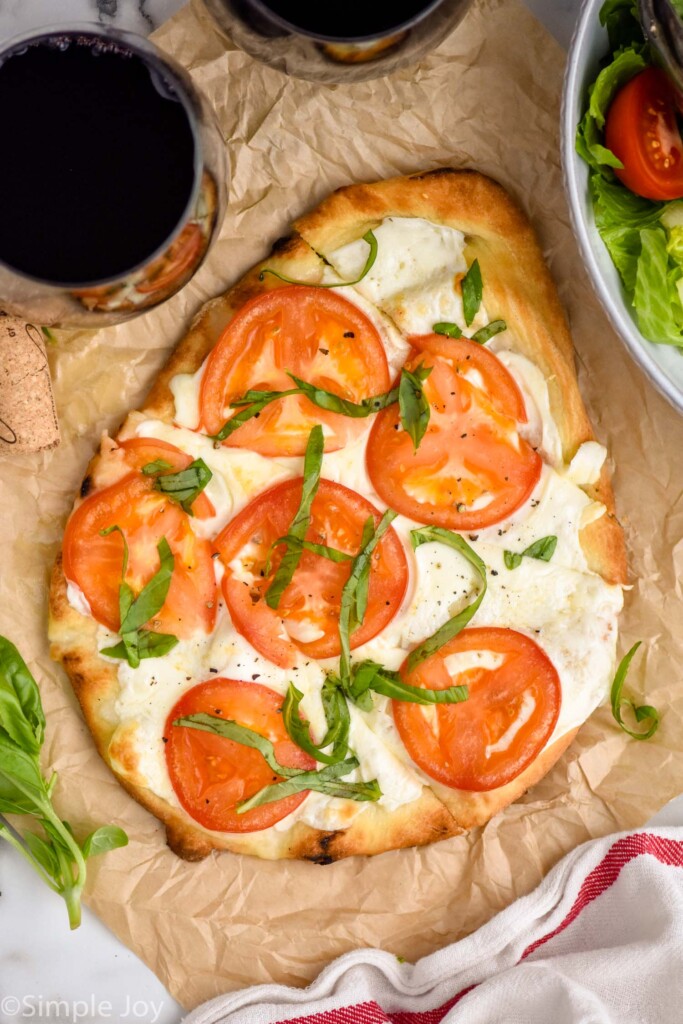 overhead of Flatbread Pizza topped with tomato slices and fresh basil. Two glasses of red wine sitting beside