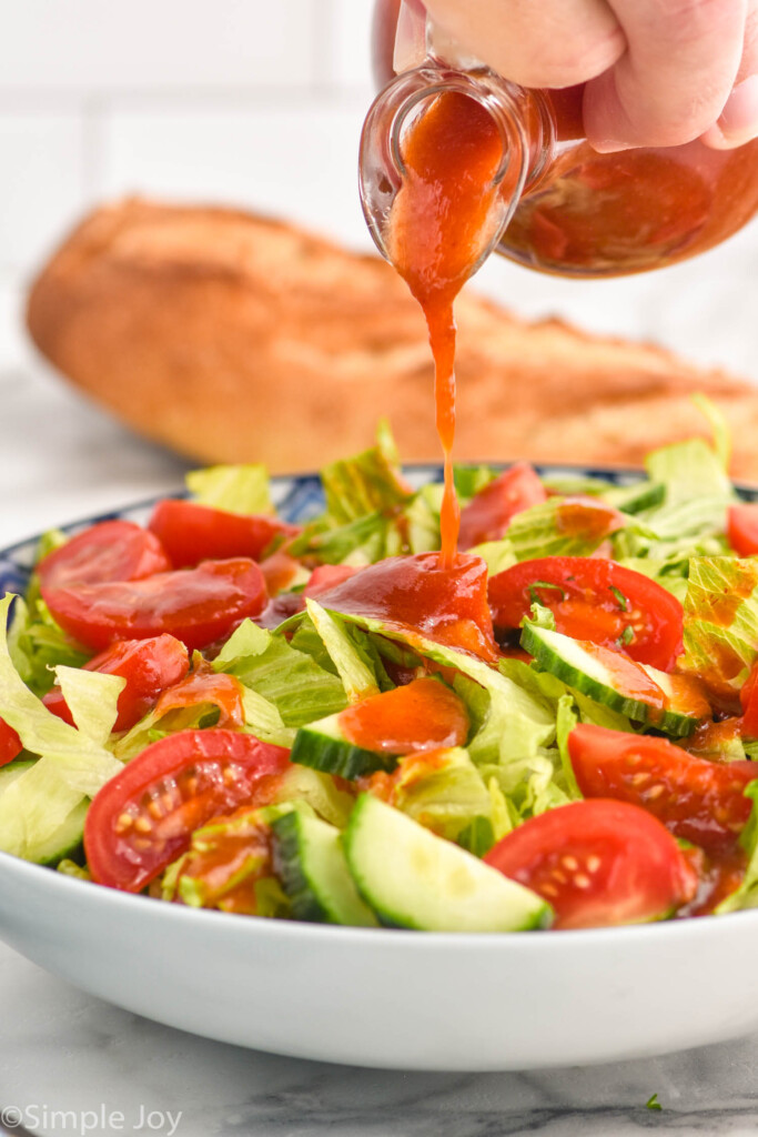 hand pouring jar of homemade French Dressing onto a bowl of lettuce salad with sliced tomatoes and cucumbers