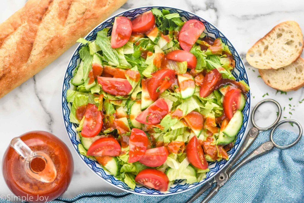 overhead of bowl of lettuce salad with sliced tomatoes, cucumbers, and homemade French Dressing. Jar of french dressing and baguette sitting beside