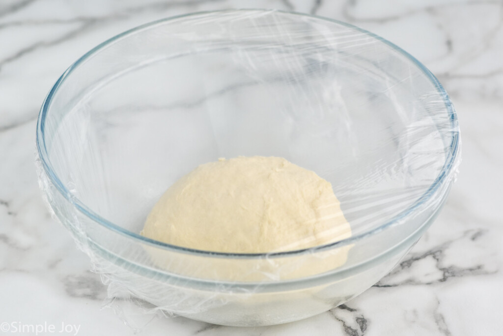 Large mixing bowl with ball of Pizza Dough covered in plastic wrap for Pizza Dough recipe.