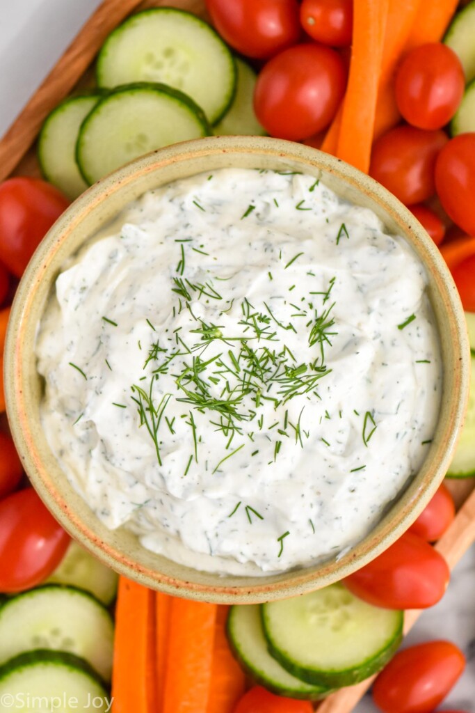 overhead of board of sliced cucumbers, carrot sticks, cherry tomatoes, and bowl of ranch dip topped with dill