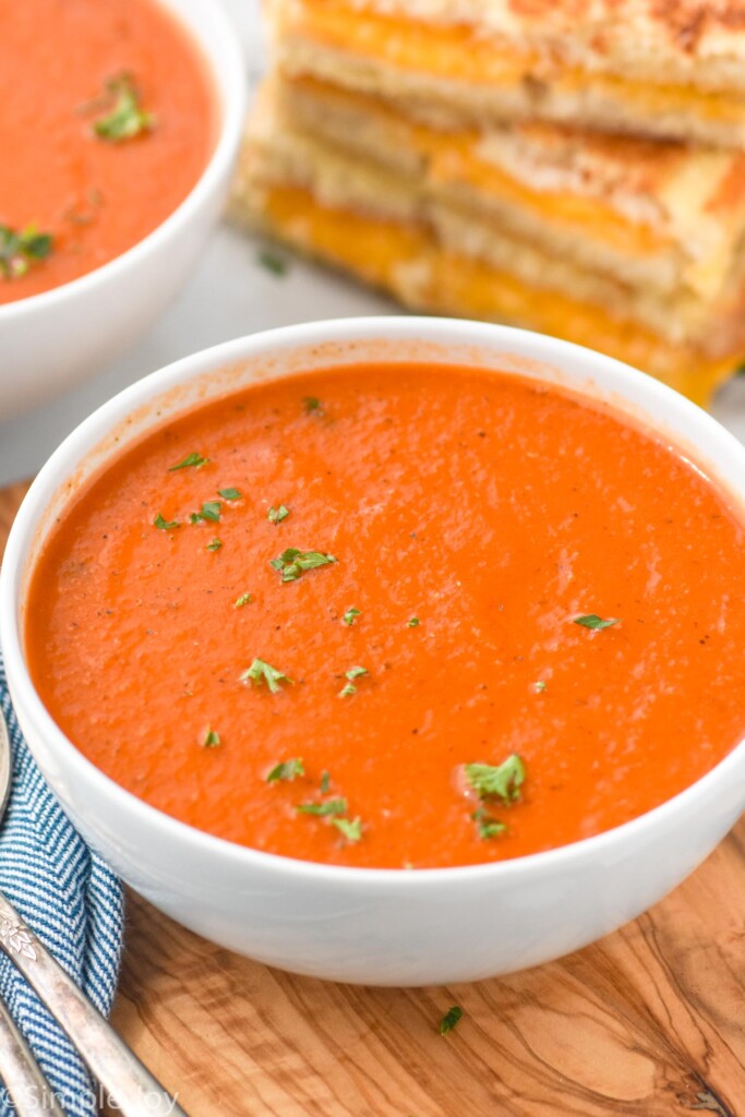 Bowl of Tomato Soup garnished with parsley. Stack of grilled cheese sandwiches beside.