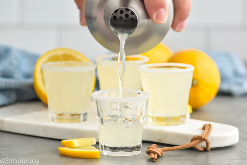 Hand pouring cocktail shaker of Lemon Drop Shot ingredients into a shot glass that is rimmed with sugar. Lemon Drop Shots, lemons, and lemon slices sitting behind