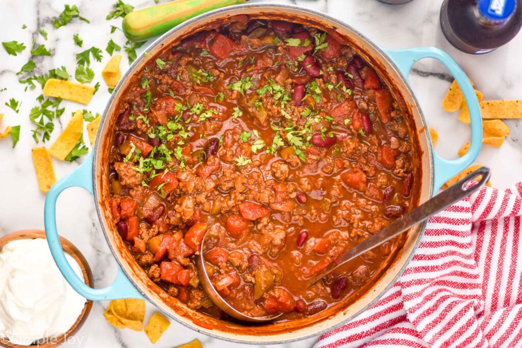 overhead of large pot of chili topped with fresh parsley and soup ladle. Corn chips, bowl of sour cream, and more fresh parsley surrounding pot.