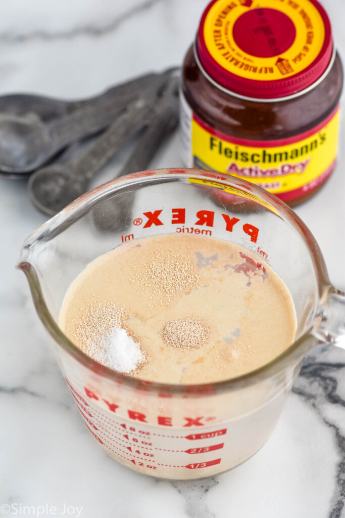 Measuring cup with water and yeast for Pizza Dough recipe, with jar of yeast and measuring spoons beside.