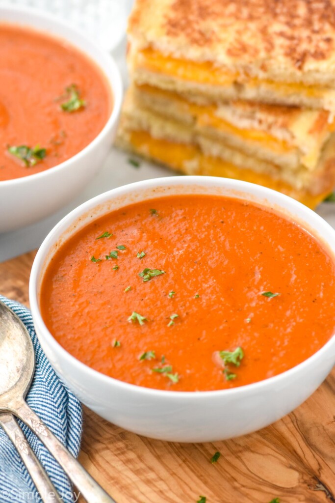 Bowl of Tomato Soup garnished with parsley. Another bowl of soup and a stack of grilled cheese sandwiches beside. Spoons beside bowls.