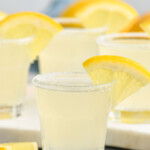 Shot glass of Lemon Drop Shot with sugared rim and slice of lemon for garnish. Lemon Drop Shots sitting in background, slices of lemon sitting beside