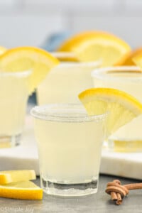 Shot glass of Lemon Drop Shot with sugared rim and slice of lemon for garnish. Lemon Drop Shots sitting in background, slices of lemon sitting beside