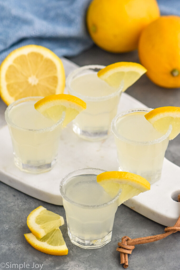 Four shot glasses of Lemon Drop Shots garnished with a slice of lemon. Fresh lemons and lemon slices sitting beside