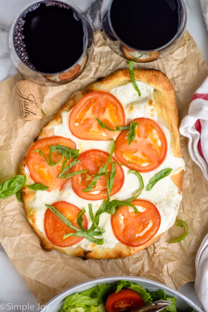 overhead of Flatbread Pizza topped with tomato slices and fresh basil. Two glasses of red wine sitting beside