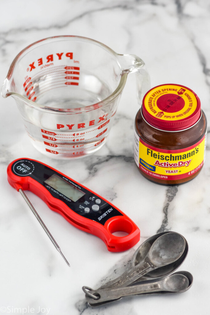 Photo of glass measuring cup of water, jar of yeast, instant read thermometer, and measuring spoons on the counter for Pizza Dough recipe.