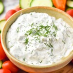 Bowl of ranch dip topped with fresh dill surrounded by carrot sticks, cucumber slices, and cherry tomatoes