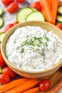 Bowl of ranch dip topped with fresh dill surrounded by carrot sticks, cucumber slices, and cherry tomatoes