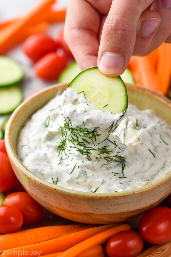 Hand holding cucumber slice dipped in ranch dip topped with fresh dill. Fresh carrot sticks, cucumber slices, and tomatoes surrounding.