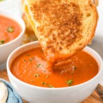 Bowl of Tomato Soup garnished with parsley with half of grilled cheese sandwich being dipped in it. Another bowl of Tomato Soup and spoons beside.