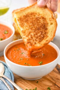 Bowl of Tomato Soup garnished with parsley with half of grilled cheese sandwich being dipped in it. Another bowl of Tomato Soup and spoons beside.