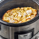 Pinterest graphic for Crockpot French Toast. Image shows a slow cooker of Crockpot French Toast dusted with powdered sugar, bowl of fresh berries and two white mugs of coffee sitting in background. Text says "Crockpot French Toast simplejoy.com"