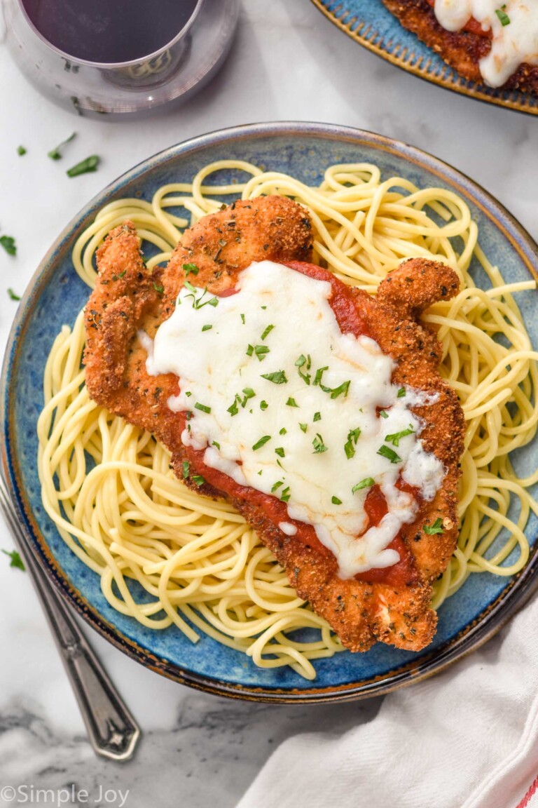 plate of spaghetti noodles topped with Chicken Parmesan, melted cheese, and fresh parsley. Fork and glass of red wine sitting beside