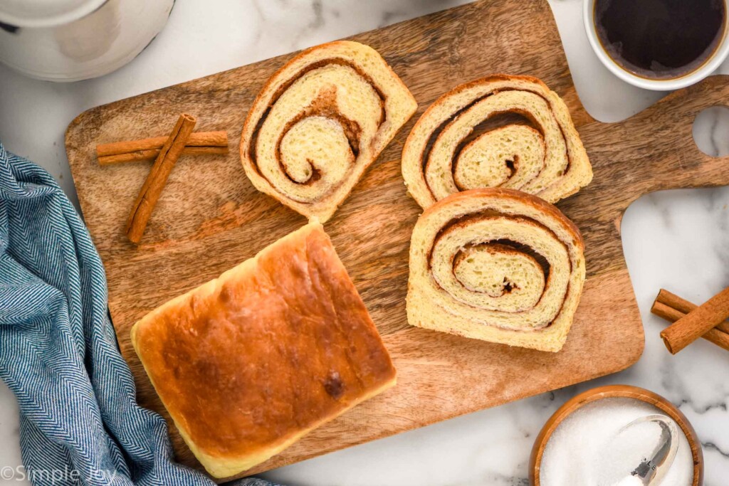 Overhead of wooden serving board with loaf of cinnamon bread, three slices of cinnamon bread, and two cinnamon sticks with mug of black coffee, cinnamon sticks, and bowl of sugar sitting beside