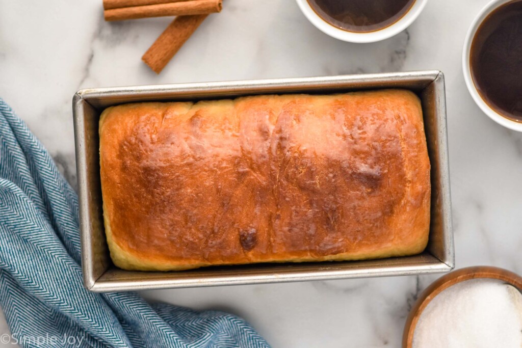 Overhead of bread pan with loaf of cinnamon bread, two mugs of coffee, cinnamon sticks, and bowl of sugar sitting beside