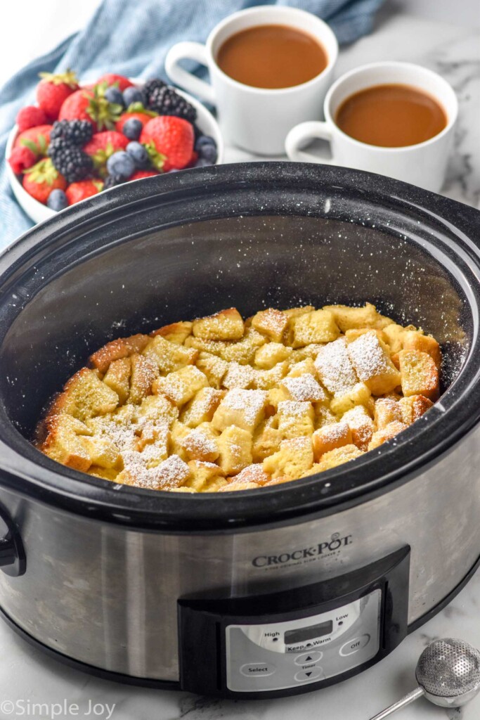 Crockpot of crockpot french toast topped with powdered sugar. Two mugs of coffee and bowl of fresh berries sitting in background