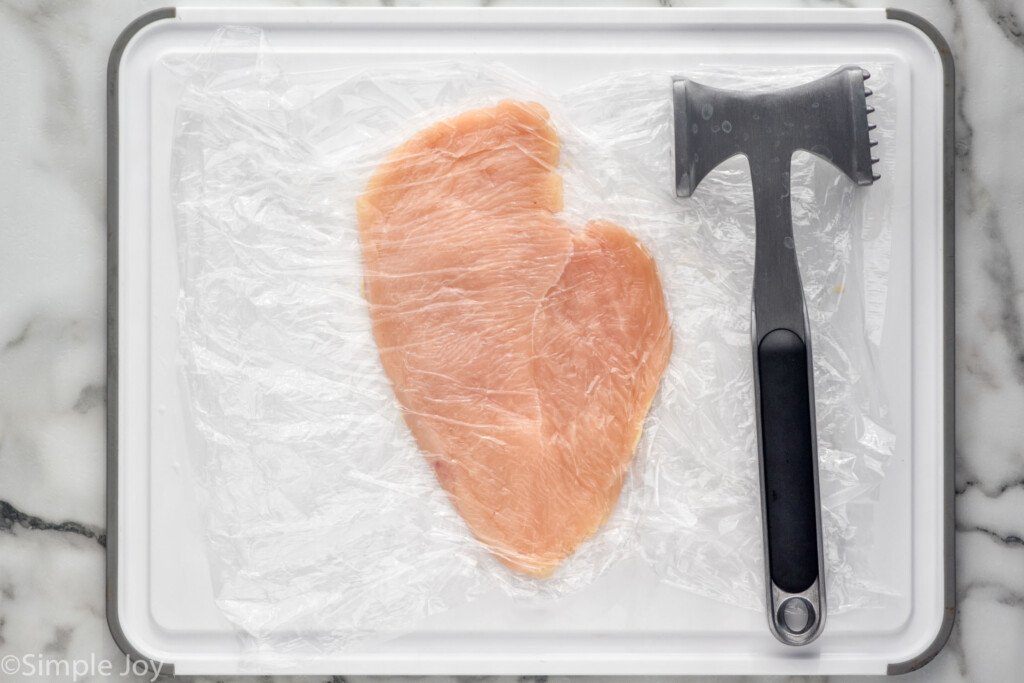 overhead of cutting board with chicken breast cutlet covered in plastic wrap with meat mallet sitting beside to make Chicken Parmesan