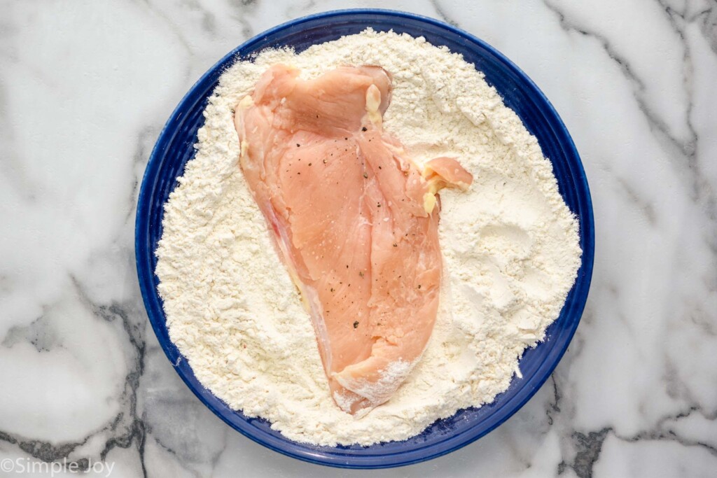 overhead of plate of flour mixture with a chicken cutlet on top to make Chicken Parmesan