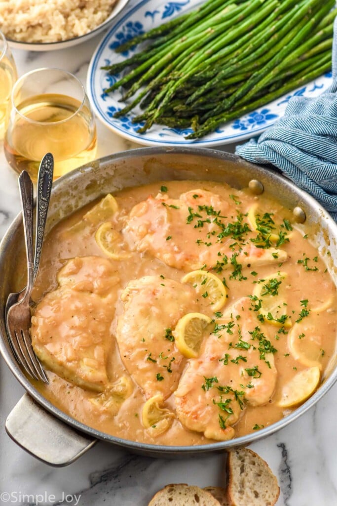 skillet of Lemon Chicken topped with lemon slices and fresh parsley. Plate of asparagus and glass of white wine sitting in background