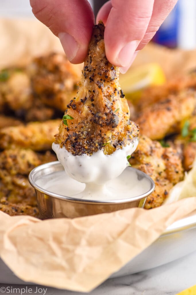 man's hand holding Lemon Pepper Wing and dipping into small cup of white dipping sauce with Lemon Pepper Wings sitting behind