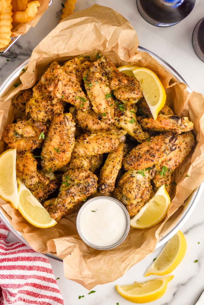 overhead of basket of lemon pepper wings with lemon slices and dipping sauce. Lemon wedges and basket of french fries sitting beside basket