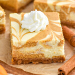 close up of Pumpkin Cheesecake Bar with dollop of whipped cream. Cinnamon sticks sitting beside and Pumpkin Cheesecake Bars sitting in background