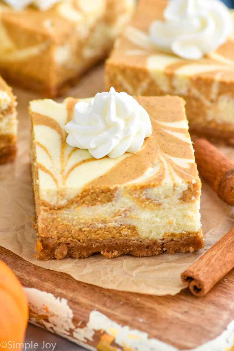 close up of Pumpkin Cheesecake Bar with dollop of whipped cream. Cinnamon sticks sitting beside and Pumpkin Cheesecake Bars sitting in background