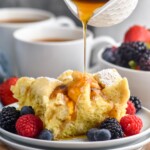 cup of syrup pouring onto a piece of crockpot french toast with powdered sugar. Fresh raspberries, blueberries, and blackberries sitting beside. Two mugs of coffee and a bowl of fruit sitting behind.