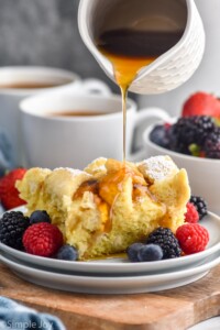 cup of syrup pouring onto a piece of crockpot french toast with powdered sugar. Fresh raspberries, blueberries, and blackberries sitting beside. Two mugs of coffee and a bowl of fruit sitting behind.