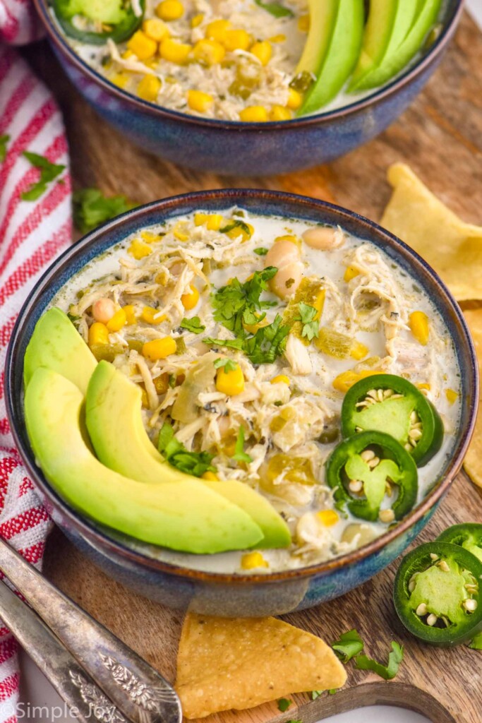 Bowl of Crockpot White Chicken Chili topped with fresh cilantro, avocado slices, and jalapeño slices. Tortilla chips and jalapeño slices sitting beside, bowl of soup in background