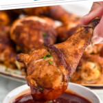 Pinterest graphic for Oven Baked Chicken Legs. Says "the best baked chicken legs simplejoy.com" Image shows hand holding baked chicken leg dipped in small bowl of bbq sauce with platter of Oven Baked Chicken Legs sitting in background