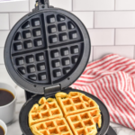 Pinterest graphic for Waffle Recipe. Text says "the best Waffle Recipe simplejoy.com" Image shows a waffle maker with homemade waffle inside, bowl of fresh berries and two mugs of black coffee sitting beside