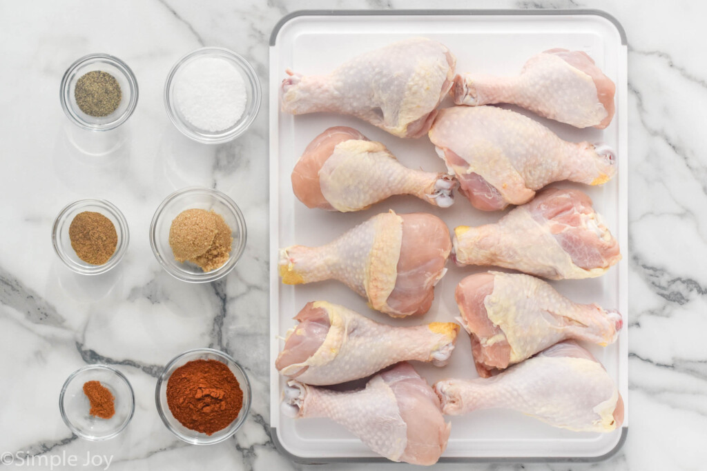 overhead of raw chicken legs on a cutting board with small bowls of seasonings sitting beside to make Oven Baked Chicken Legs