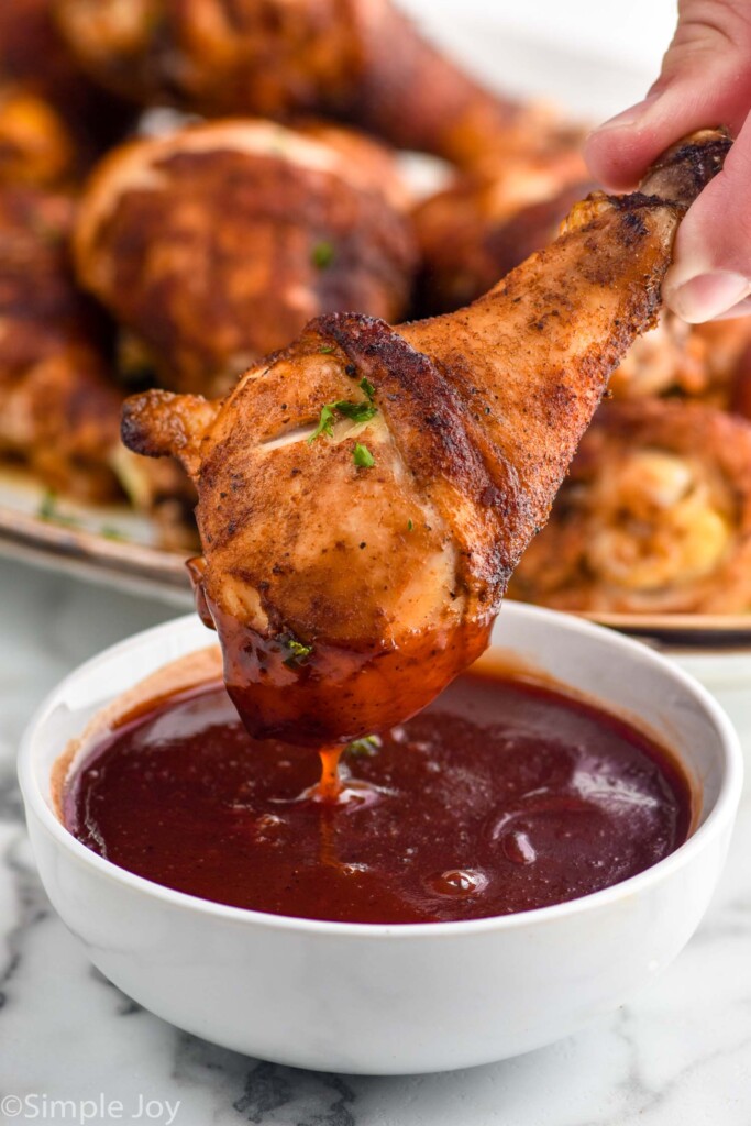 hand holding Oven Baked Chicken Leg dipped in small bowl of bbq sauce with platter of Oven Baked Chicken Legs sitting in background