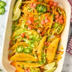 overhead of baking dish of baked tacos topped with melted cheese, shredded lettuce, diced tomatoes, diced Jalapeños, and lime wedges