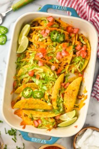 overhead of baking dish of baked tacos topped with melted cheese, shredded lettuce, diced tomatoes, diced Jalapeños, and lime wedges