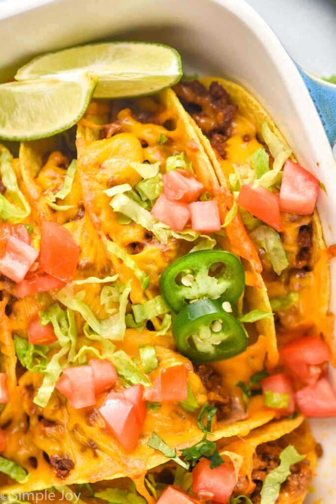 Close up of baked tacos in a baking dish topped with melted cheese, diced tomatoes, shredded lettuce, lime wedges, and diced jalapeños