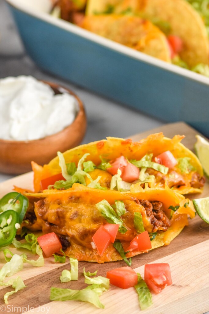 two baked tacos topped with melted cheese, diced tomatoes, shredded lettuce, and diced Jalapeños with bowl of sour cream and casserole dish of baked tacos sitting in background