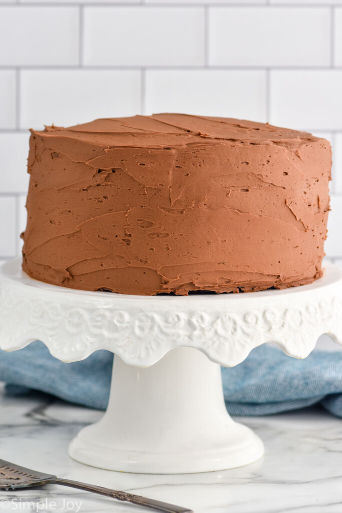 Frosted chocolate cake on a white cake stand with spatula for serving sitting beside