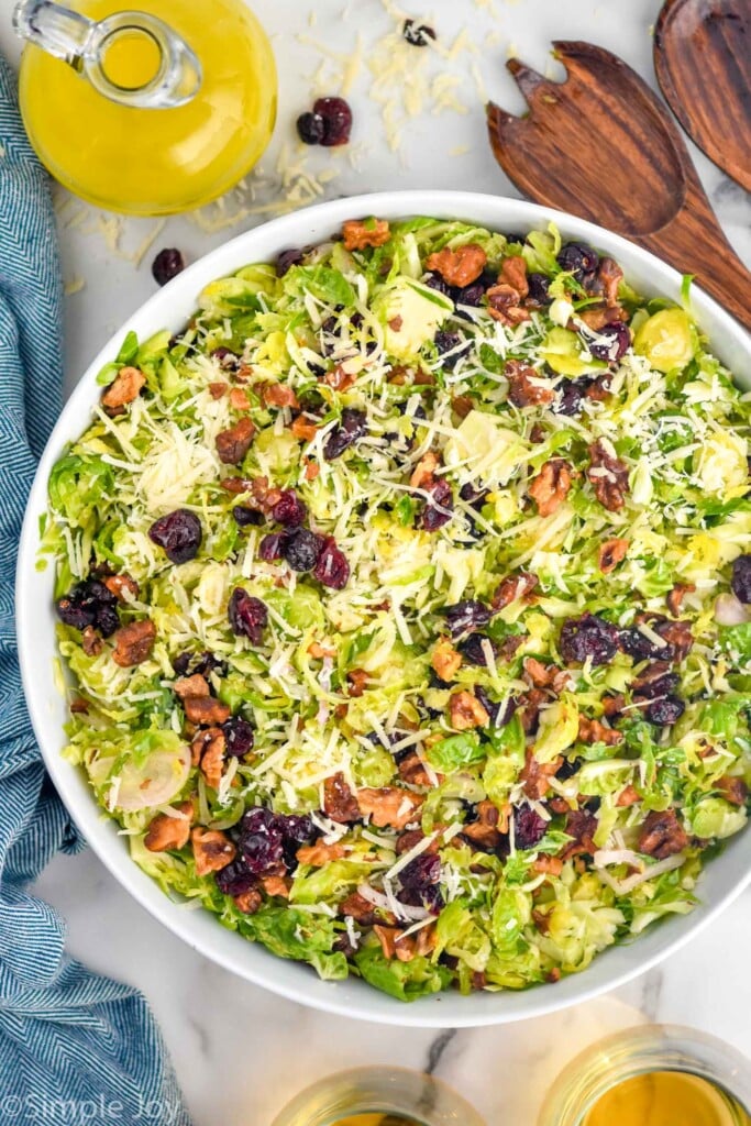 overhead of bowl of Shaved Brussels Sprout Salad topped with shredded parmesan cheese. Wooden utensils and jar of dressing sitting beside