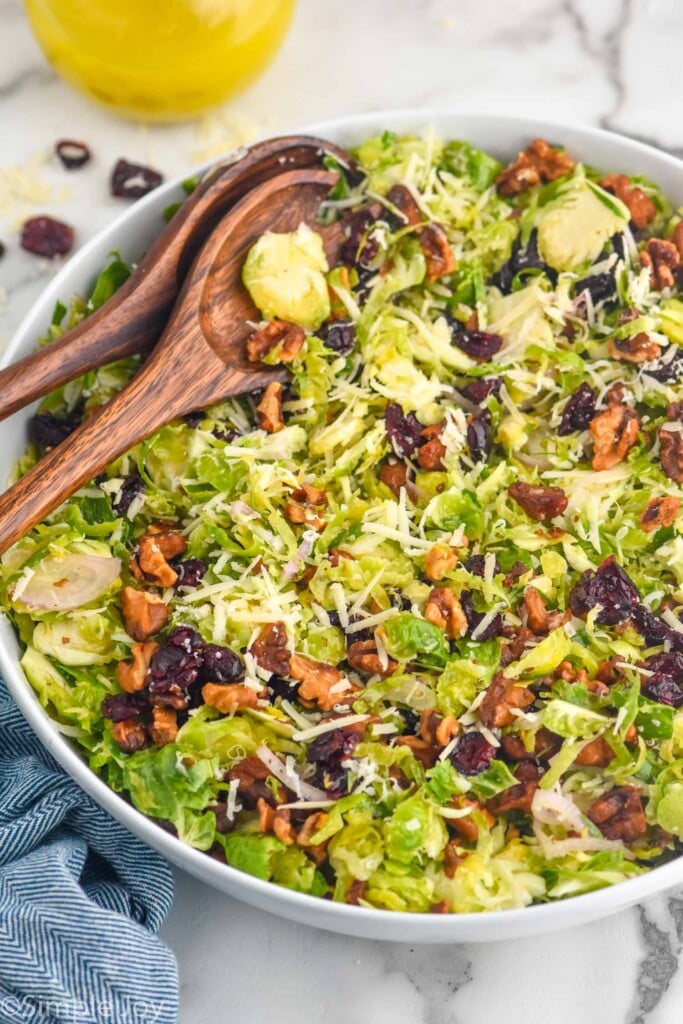 bowl of Shaved Brussels Sprout Salad topped with shredded parmesan cheese, walnuts, and dried cranberries with two wooden spoons for serving.