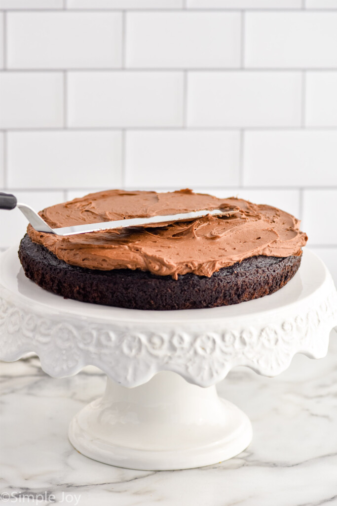 Single layer of Chocolate Cake sitting on a cake stand with dollop of chocolate frosting and spatula on top