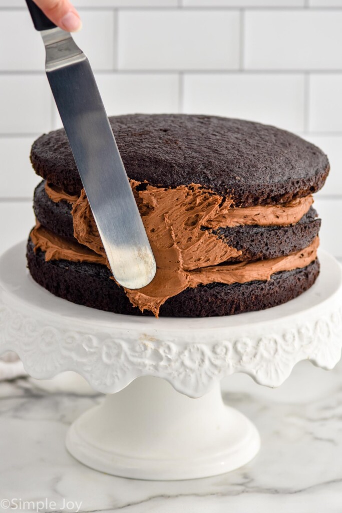 Layered Chocolate Cake sitting on cake plate with spatula frosting the sides