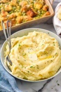 bowl of crockpot mashed potatoes topped with butter and fresh parsley with two spoons for serving. Casserole dish of brussels sprouts casserole sitting in background