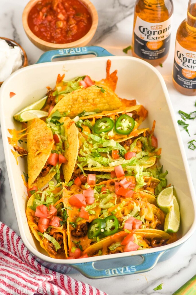 baking dish of Baked Tacos topped with diced tomatoes, shredded lettuce, lime wedges, and diced Jalapeños. Bowl of salsa and two bottles of beer sitting behind